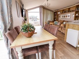 A dining table with a flower basket and four chairs in a room with sofas shelves and a large window at Hazel Park Lodge HP4 in Rhyl