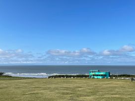A grassy field near a beach with a teal double-decker bus and a person carrying a surfboard near the shore at The Links 40 An apartment with spectacular sea views in Porthcawl