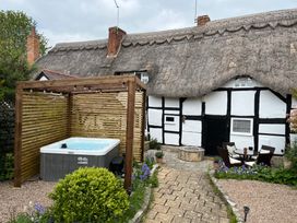 An outdoor area with a hot tub under a wooden pergola next to a thatched roof cottage with white walls and black timber framing at Church View in Welford-On-Avon