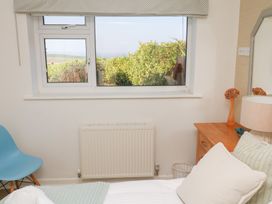A bedroom with a window overlooking greenery a blue chair a wooden bedside table with a lamp and a flower vase at Coastmans Nest in Mawgan Porth