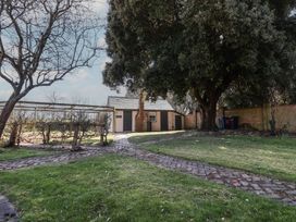 An outdoor garden area with a stone path leading to a small building with a chimney and large trees at The Woodshed in Tendring near Weeley