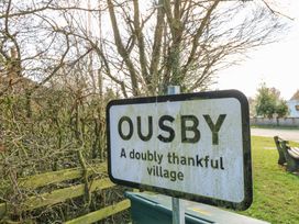 A village sign with the text Ousby A doubly thankful village near trees and a bench at Rayson Hall Barns in Ousby