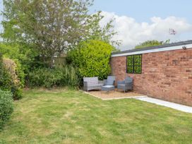 A garden area with grass, shrubs, and outdoor wicker chairs and table next to a brick wall with a window at Little Lodge in Dersingham