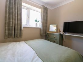 A bedroom with a bed covered by a blanket a window with plaid curtains a green chest of drawers with a mirror and a wooden duck and a wall-mounted TV at 6 Barleycroft in Hemsby