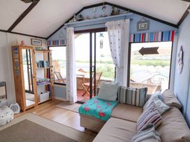 A living room with a beige sectional sofa with striped pillows and flamingo blanket wooden bookshelf with games and a mirror sliding glass doors with white curtains leading to a balcony with wooden chairs and table at Captain's Cabin in Clarach Bay near Aberystwyth
