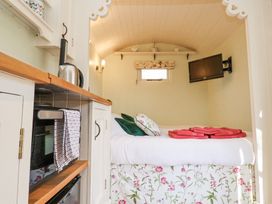 A small room with a bed, pillows, red towels, a wall-mounted TV, and a kitchenette with a kettle and oven at Woodlands Retreat Shepherds Hut Brundish near Laxfield