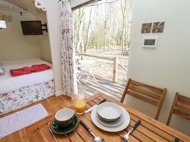 A dining area with a wooden table set with plates cups and cutlery next to a bed with towels and a TV and an open door leading to a wooden deck with outdoor furniture and trees at Woodlands Retreat Shepherd's Hut Brundish near Laxfield