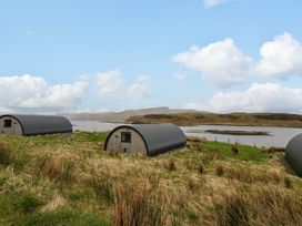 Three small curved wooden cabins near a body of water with grassy land and hills in the background at WildLuing in Cullipool Isle Of Luing