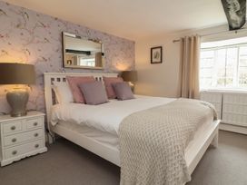 A bedroom with a white bed frame, pink and purple pillows, two lamps on nightstands, a window with beige curtains and floral wallpaper behind the bed at Dragon House in Bilbrook near Minehead