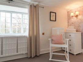 A room with a white rocking chair with a pink cushion next to a white chest of drawers with folded towels on top and a window with beige curtains at Dragon House in Bilbrook near Minehead