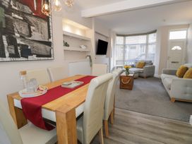 A dining area with a wooden table and cream chairs next to a living room with grey sofas and a wooden chest at 21 Taliesin Street in Llandudno
