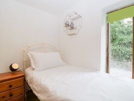 A single bed with white bedding next to a wooden nightstand with a small lamp and a window with a green blind showing outside greenery at Mistletoe Cottage in Kemeys Commander near Usk
