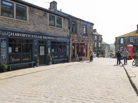 A street with shops including Haworth Steam Brewing Co and Pave Cheesemonger with people walking and a red phone booth in Haworth