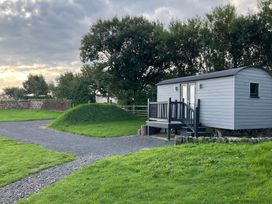 A small gray wooden cabin with stairs and a railing on a grassy area with trees and a stone wall at Little Acre in Oxenhope