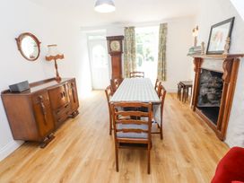 A dining room with a wooden table covered by a tablecloth surrounded by six chairs next to a mantelpiece with decorations and a sideboard with a lamp and mirror at Llys Emrys in Beddgelert