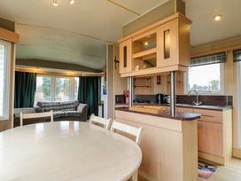 A dining area with a round wooden table and chairs next to a kitchen with cabinets a countertop and a sink and a living area with a sofa and windows at Beatrice's Static Caravan in Spalding