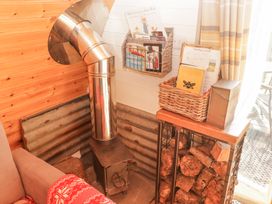 A small wood burning stove with a metal pipe surrounded by wood logs a basket with papers and a wire basket with books on a white wooden wall at Cabanau Bach in Bedwas