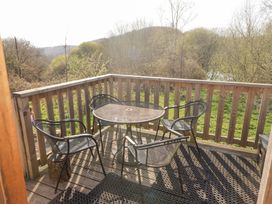 A wooden balcony with a round metal table and four metal chairs overlooking trees and grassy hills at Cabanau Bach in Bedwas