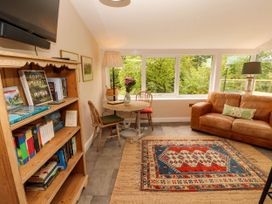 A living room with a wooden bookshelf a round table with two chairs a brown leather sofa and large windows overlooking trees at Kirkland Howe Cottage in Arlecdon