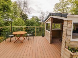 An outdoor wooden deck with a round table two chairs glass railing and a small wooden cabin surrounded by trees at Kirkland Howe Cottage in Arlecdon