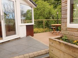 A wooden deck with a round table and chairs next to a house with large windows and a planter box with plants at Kirkland Howe Cottage in Arlecdon