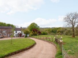 A curved driveway leading to a house with stone and white walls next to a grassy field with sheep and trees at Kirkland Howe Cottage in Arlecdon