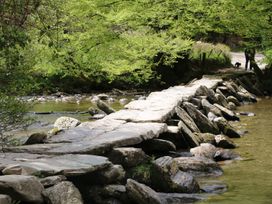 A stone footbridge over a shallow river with green trees and a person with a dog in the background at 1 The Mews in Porlock
