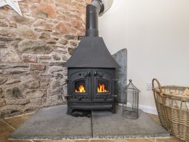 A wood-burning stove with fire inside set against a stone wall with a basket of firewood and a metal lantern beside it at 1 The Mews in Porlock
