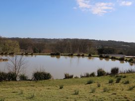 A pond surrounded by grass and bushes with trees and hills in the background at Tench Hut Hadlow Down near Crowborough