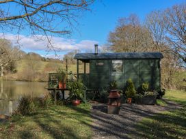 A green wooden cabin with plants and a fire pit near a pond at Roach Hut in Hadlow Down near Crowborough