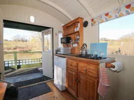 A compact kitchen with wooden cabinets a gas stove a kettle microwave and a fridge next to an open door showing a view of a pond and trees at Roach Hut in Hadlow Down near Crowborough
