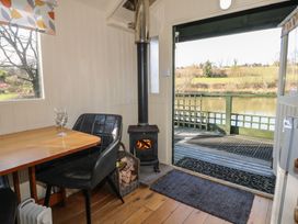A small indoor dining area with a table and two black chairs next to a wood burning stove and an open door leading to a deck overlooking a body of water at Roach Hut in Hadlow Down near Crowborough