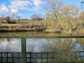 A view of a pond with trees and a grassy field in the background taken from behind a wooden fence at Roach Hut in Hadlow Down near Crowborough
