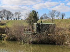 A wooden cabin with a small deck by a pond with trees and grassy hills in the background at Carp Hut in Hadlow Down near Crowborough