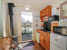 A small kitchen area with wooden cabinets a stove tea kettles a microwave and a wood burning stove next to an open door leading to a patio with a table and chairs by water at Carp Hut in Hadlow Down near Crowborough