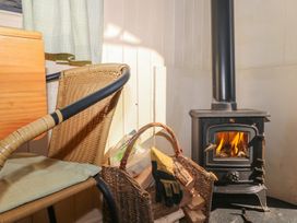 A wood burning stove with a fire inside next to a wicker basket filled with firewood and gloves and a chair with a cushion at Carp Hut in Hadlow Down near Crowborough