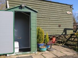 A small green outdoor shed with an open door revealing a white portable toilet nearby plants in blue pots on a stone patio at Carp Hut in Hadlow Down near Crowborough