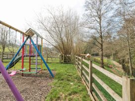 A fenced grassy area with a colorful swingset and slide next to trees and a small creek at Bell tent 3 in Camerton near Timsbury