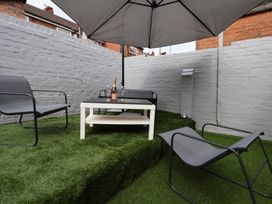 An outdoor seating area with a white table holding a bottle and glasses under a large umbrella and three gray chairs on artificial grass at Anfield Stadium Short Stay and Free Parking in Liverpool