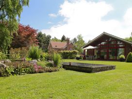 A garden with a lawn, flower beds, and two houses with large windows and patio furniture at Studio Cottage in Surlingham near Poringland