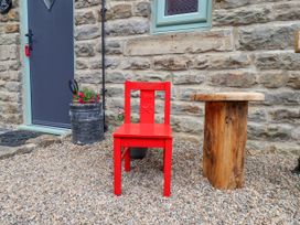 A red chair and a wooden stump table on gravel outside a stone wall with a gray door and small window at Wayside Farm Dairy Cottage in Cloughton near Burniston