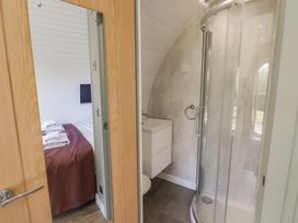 A bathroom with a curved glass shower enclosure and a white sink next to a doorway showing a bedroom with a bed covered by a brown blanket at The Rowan Pod in Cowbridge