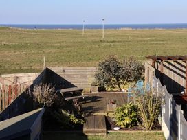A backyard with wooden deck, picnic table, blue chairs, plants, fence, grassy field, and sea in the background at 48 Waxholme Road in Withernsea