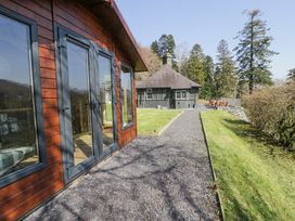 A gravel pathway between a wooden building and a grassy area leading to a small dark-colored house with trees in the background at Bwthyn y Coediwr The Woodcutters Cottage in Tan Y Bwlch Maentwrog