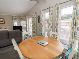 A dining area with a round wooden table and white chairs with cushions near windows with patterned curtains at Lodge 2 in Auchterarder