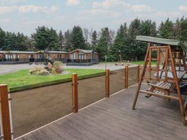 A wooden swing on a deck overlooking a garden with decorative plants and wooden lodges in the background at Lodge 2 in Auchterarder