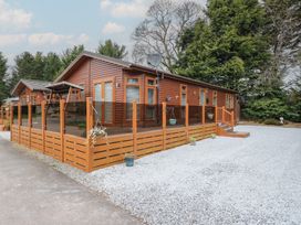 A wooden cabin with a fenced deck and gravel driveway surrounded by trees at Lodge 2 in Auchterarder