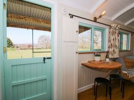 A room with a turquoise half door opening to a field an indoor wooden table with two stools a wicker chair and windows with floral curtains at Gilberry Shepherds Hut Wall under Heywood near Church Stretton