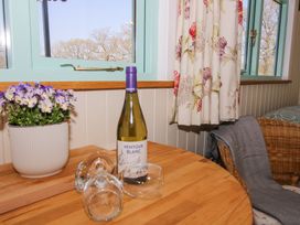A wooden table with a bottle of Ventoux Blanc wine, three wine glasses, a potted purple flower, floral curtains, and a wicker chair with a gray blanket at Gilberry Shepherds Hut Wall under Heywood near Church Stretton