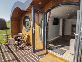 An outdoor wooden deck with a round table and four chairs leading to an open door showing a living area with a wall-mounted TV and kitchen at The Hazel Pod in Cowbridge